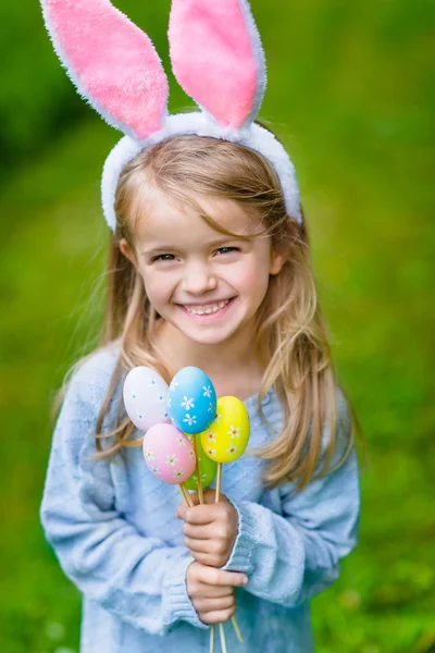 Bella bambina sorridente con lunghi capelli biondi che indossa orecchie di coniglio rosa o coniglio e abito blu a maglia e che tiene mazzo di uova colorate dipinte nella giornata di sole nel parco primaverile. Celebrazioni pasquali — Foto Stock