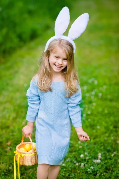 Glimlachend meisje met lang blond haar dragen wit konijn bunny oren en blauwe gebreide jurk en het houden van rieten mand met gele eieren en lint op zonnige dag in Spring Park. PaaS vieringen — Stockfoto