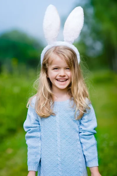 Ritratto all'aperto di una bella bambina sorridente con lunghi capelli biondi che indossa orecchie bianche di coniglio o coniglio e un vestito blu a maglia nella giornata di sole nel parco primaverile. Celebrazioni pasquali — Foto Stock