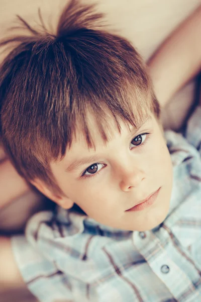 Closeup portrait of pensive little boy with brown eyes wearing checkered shirt lying on floor and looking at camera. Happy childhood concept, selective focus on eyes, top view, instagram filters — Stock Photo, Image