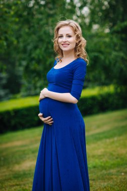 Adorable smiling young pregnant woman in blue dress with long blond curly hair holding her belly in summer park on rainy day. Pregnancy and femininity concept. Waterdrops on blue dress