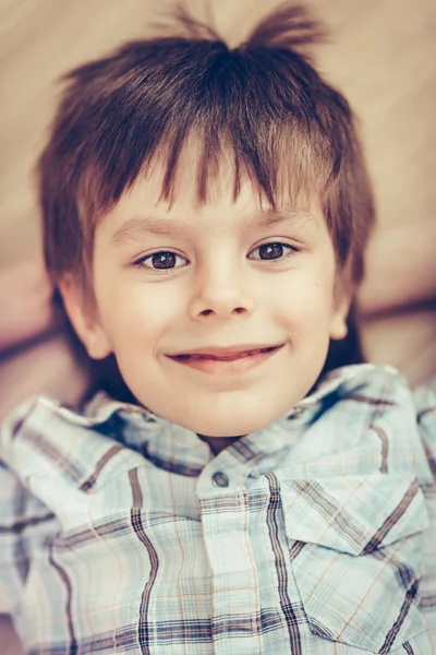 Closeup portrait of smiling little boy with brown eyes wearing checkered shirt lying on floor and looking at camera. Happy childhood concept, selective focus on eyes, top view, instagram filters — Stock Photo, Image