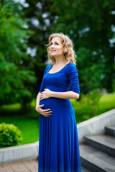 Belle jeune femme enceinte en robe bleue avec de longs cheveux bouclés blonds tenant son ventre, souriant et regardant le ciel dans le parc d'été le jour de pluie. Concept de grossesse et de féminité. Gouttes d'eau sur robe — Photo