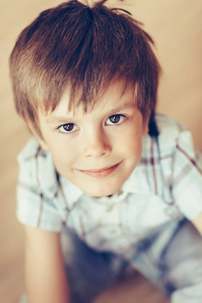 Closeup portrait of handsome smiling little boy with brown eyes wearing checkered shirt sitting on floor looking at camera. Happy childhood concept, selective focus on eyes, top view, instagram filter — Stock Photo, Image