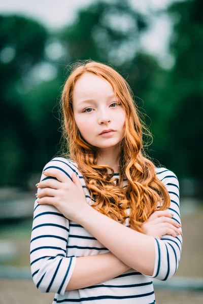 Lovely red-haired girl hugging herself and looking at camera. Beautiful young woman with red hair and green eyes in summer park. Sad teenage girl feeling herself lonely. Solitude, loneliness concept