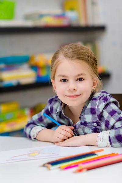 Preciosa niña sonriente con el pelo rubio sentado en la mesa blanca con lápices multicolores y mirando a la cámara — Foto de Stock