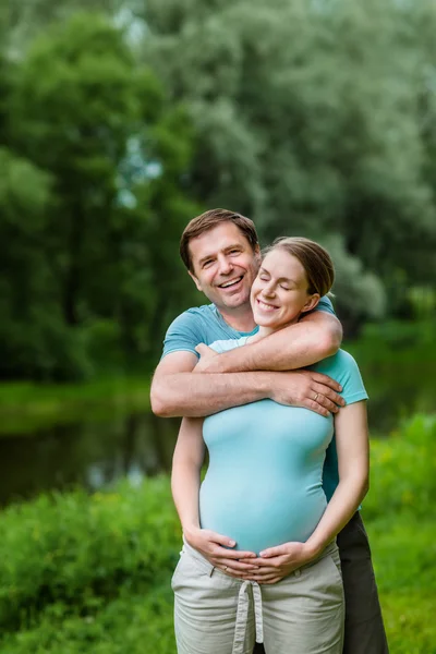 Felice uomo bello sorridente abbracciando la sua bella giovane donna incinta che tiene la pancia nel parco estivo. Concetto di famiglia felice e gravidanza. Festa della mamma — Foto Stock