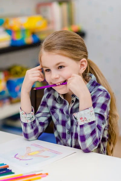 Petite fille drôle avec les cheveux blonds assis à la table blanche et tenant crayon violet dans sa bouche — Photo