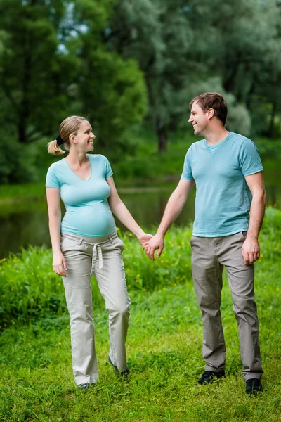 Adorable joven embarazada y feliz hombre guapo sonriendo, tomándose de las manos y mirándose en el parque de verano. Concepto de familia feliz y embarazo. Día de la Madre —  Fotos de Stock