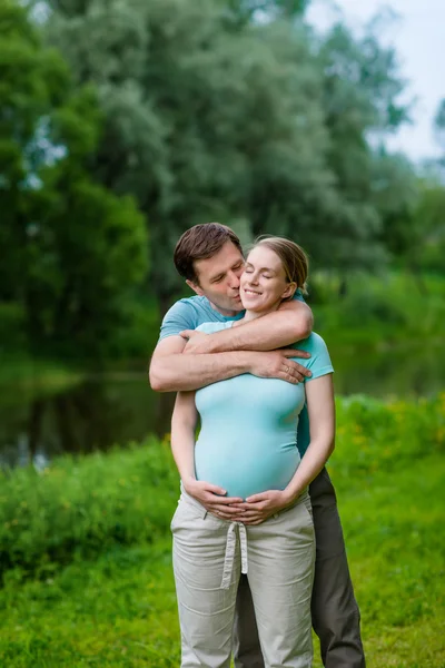 Glücklich lächelnder schöner Mann umarmt und küsst seine schöne junge schwangere Frau, die ihren Bauch im Sommerpark hält. glückliche Familie und Schwangerschaft Konzept. Muttertag — Stockfoto