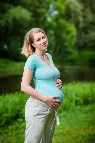 Beautiful smiling young pregnant woman wearing turquoise t-shirt and beige pants with blond hair holding her belly in summer park. Pregnancy and femininity concept. Mother's Day Royalty Free Stock Photos