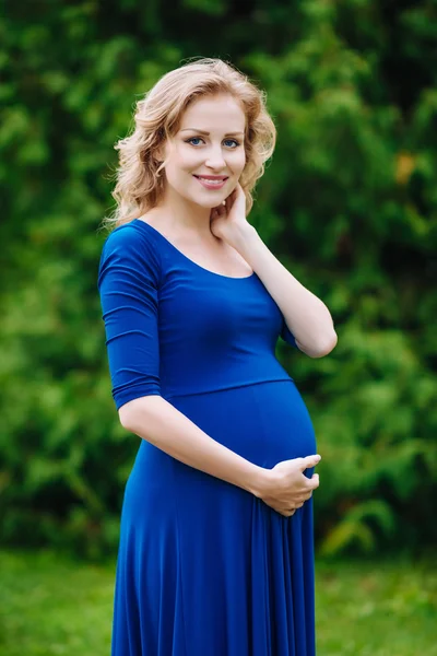 Porträt einer hübsch lächelnden jungen Schwangeren in blauem Kleid mit langen blonden lockigen Haaren, die ihren Bauch hält und im Sommerpark in die Kamera blickt. Schwangerschaft und Weiblichkeit. Muttertag — Stockfoto