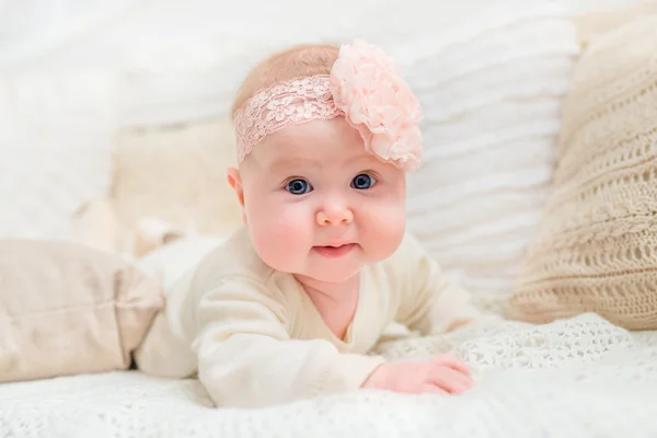 Smiling baby girl with chubby cheeks and big blue eyes wearing white clothes  and pink band with flower lying on bed and looking at camera. Babyhood and childhood concept — Stock Photo, Image