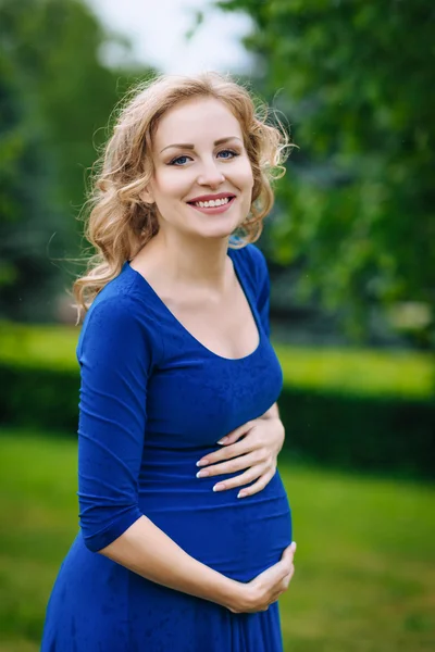 Retrato de la encantadora joven embarazada sonriente en vestido azul con el pelo largo y rubio rizado sosteniendo su vientre y mirando a la cámara en el parque de verano. Concepto de embarazo y feminidad. Día de la Madre Imágenes de stock libres de derechos