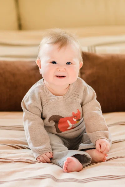 Smějící se malé dítě s blond vlasy a modré oči, nosit pletený svetr sedí na pohovce a dotýká její nohu. Šťastné dětství — Stock fotografie