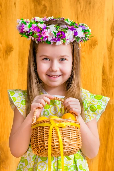 Portrait of adorable smiling little girl with long blond hair wearing floral head wreath and holding wicker basket with yellow eggs and ribbon. Easter celebrations. Wooden background. Studio portrait — Stock Photo, Image