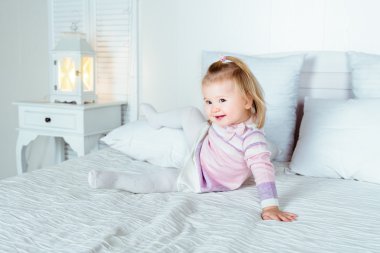 Yatak yatak odasında oynamaya komik ve sevimli sarışın küçük gülümseyen kız. Beyaz iç, büyük yatak, komidin ve gece lambası.