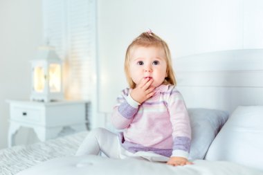 Funny amazed blond little girl sitting on bed in bedroom. White interior, big bed, bedside table and night lamp. Girl touches her mouth by hand