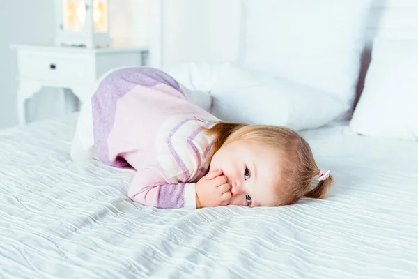 Cute little blonde girl on hands and knees on white bed in bedroom. White interior, big bed, bedside table and night lamp — Stock Photo, Image