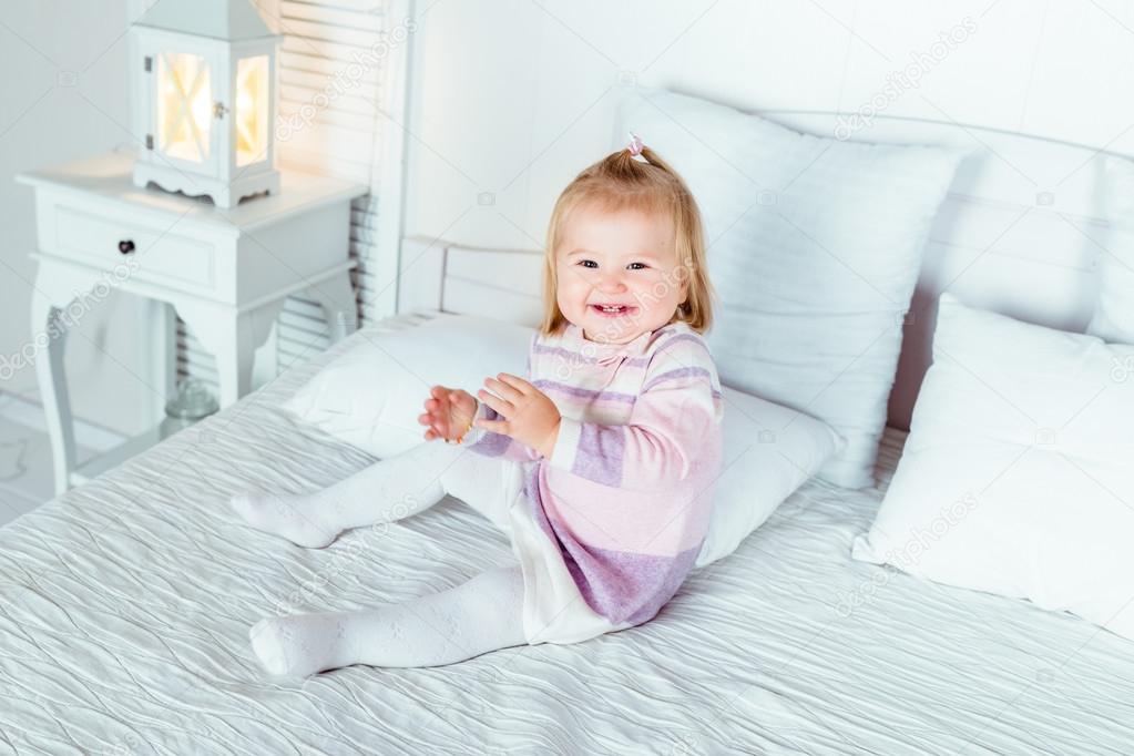 Funny and cute blond little laughing girl playing on bed in bedroom. White interior, big bed, bedside table and night lamp