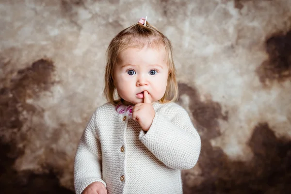 Close-up portrait of cute blond little girl with big grey eyes and plump cheeks keeping her finger in her mouth. Studio portrait on grunge background — Stock Photo, Image