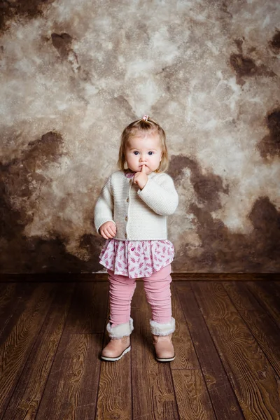 Cute blond little girl with big grey eyes and plump cheeks staying and keeping her finger in her mouth. Studio portrait on grunge background — Stock Photo, Image