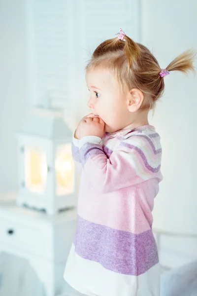 Amazed blond little girl with ponytail staying on bed and keeping her hands in her mouth. White interior, bedroom, night lamp — Stock Photo, Image