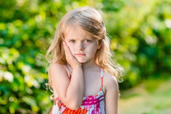 Ragazzina triste con lunghi capelli biondi che soffrono di mal di denti. Giornata estiva soleggiata in un bellissimo parco verde Immagine Stock