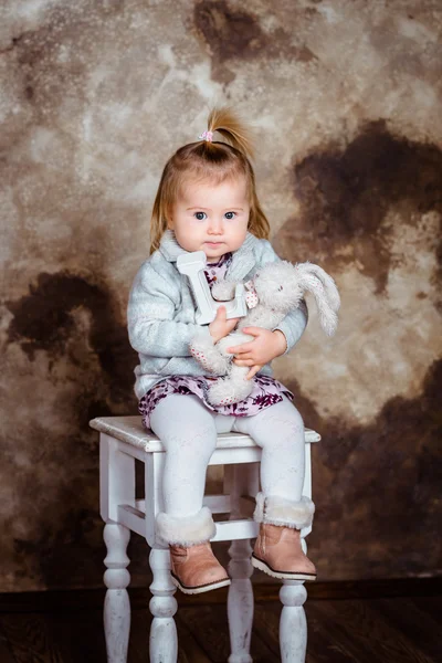 Söt blond liten flicka sitter på vit stol och hålla hennes leksaker. Studio porträtt på brun grunge bakgrund — Stockfoto