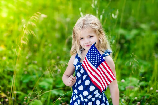 Söta leende liten flicka med långt lockigt blont hår håller en amerikansk flagga på solig dag i sommar park. Självständighetsdagen, flagga dag koncept — Stockfoto