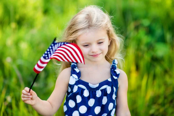 Hübsches, nachdenkliches kleines Mädchen mit langen blonden Locken, das eine amerikanische Flagge in der Hand hält und an einem sonnigen Tag im Sommerpark lächelt. Unabhängigkeitstag, Flaggentag-Konzept — Stockfoto