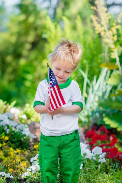 Smiling little boy with blond hair holding american flag and looking at it in sunny park or garden on summer day. Portrait of child on blurred background. Independence Day, Flag Day concept — Stock Photo, Image