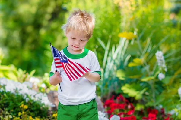 Cute pensive little boy with blond hair holding american flag and looking at it in sunny park or garden on summer day. Portrait of child on blurred background. Independence Day, Flag Day concept — Stock Photo, Image