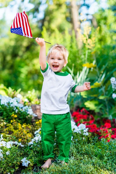 Босоногий маленький мальчик со светлыми волосами смеется и машет американским флагом в солнечном парке или саду в летний день. Портрет ребенка на размытом фоне. День независимости, концепция Дня флага — стоковое фото