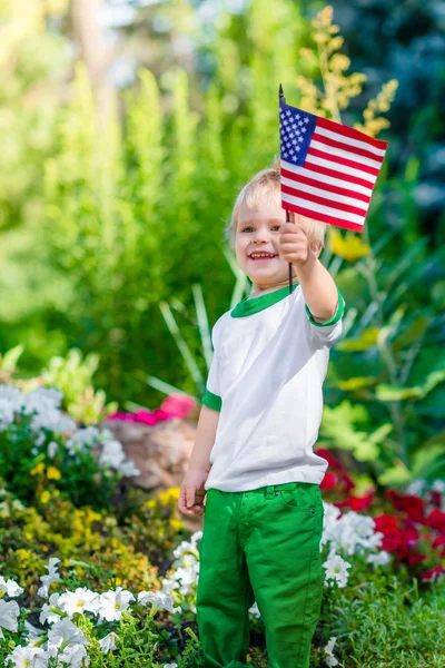 Glimlachend blond jongetje houden van Amerikaanse vlag en zwaaien het in zonnige park of tuin op zomerdag. Portret van kind op onscherpe achtergrond. Independence Day, dag van de vlag concept — Stockfoto