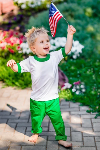 Funny laughing little boy with blond hair holding american flag and waving it in sunny park or garden on summer day. Portrait of child on blurred background. Independence Day, Flag Day concept — Stock Photo, Image