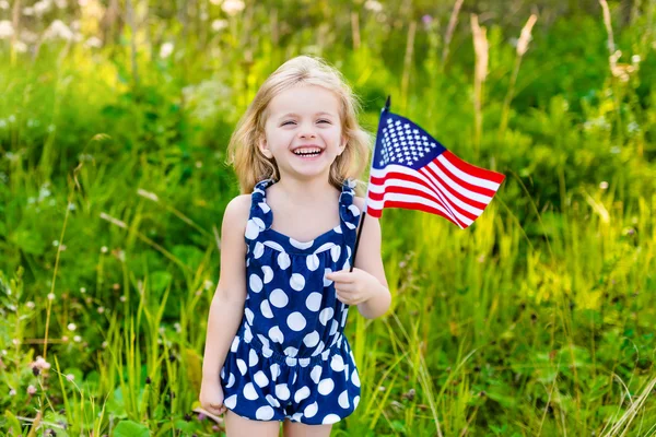Śliczną dziewczynkę z długie kręcone blond włosy z amerykańską flagę w ręce śmiejąc się w słoneczny dzień w lecie parku. Święto Niepodległości, dzień flagi koncepcja — Zdjęcie stockowe