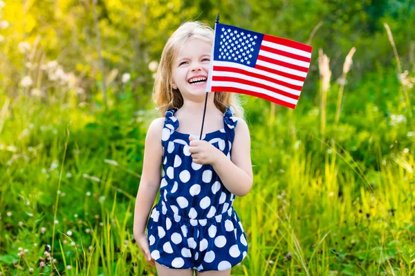 Rolig liten flicka med långt lockigt blont hår håller en amerikansk flagga, vinka det och skratta på solig dag på sommaren park. Självständighetsdagen, flagga dag koncept — Stockfoto