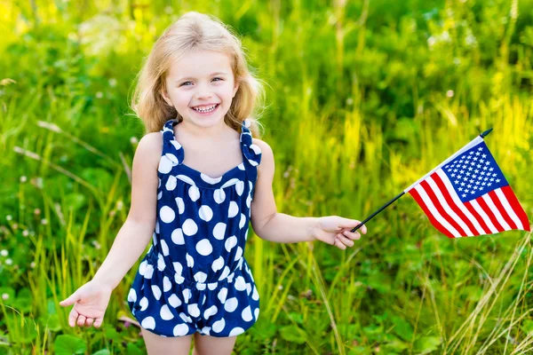 Leende liten flicka med långt lockigt blont hår med amerikanska flaggan och vinkar det, utomhus porträtt på solig dag i sommar park. Självständighetsdagen, flagga dag koncept — Stockfoto