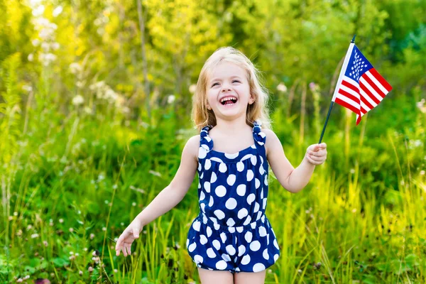 Skrattande blond liten flicka med långt lockigt hår håller amerikanska flaggan och vinkar det, utomhus porträtt på solig dag i sommar park. Självständighetsdagen, flagga dag koncept — Stockfoto