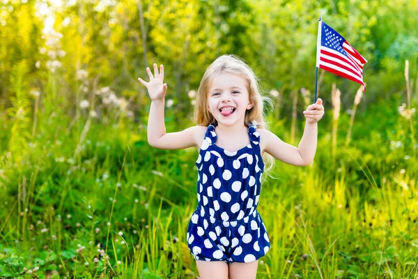 Rolig liten flicka med långt lockigt blont hår lägger ut sin tunga och vifta med amerikanska flaggan, utomhus porträtt på solig dag i sommar park. Självständighetsdagen, flagga dag koncept — Stockfoto