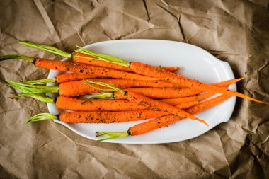 Oven-baked baby carrots sprinkled with fresh-grounded black pepper on white plate on craft paper. Healthy vegetarian snack, delicious vegan food clipart