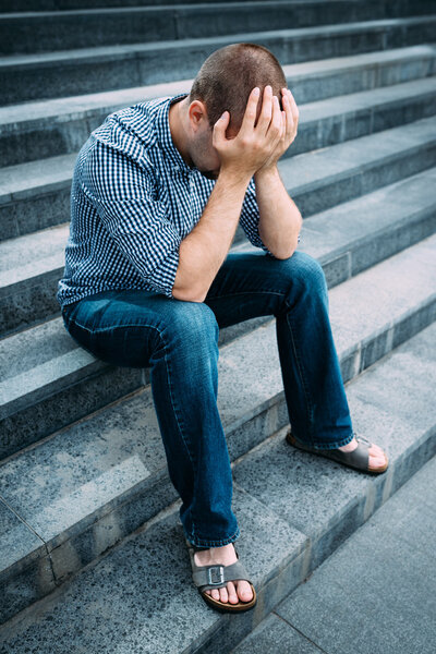 Sad young man covering his face with hands sitting on stairs of big building. Feelings of sadness, despair and tragedy