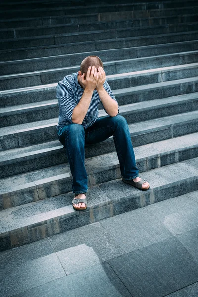 Outdoor Portret van ontredderde jonge man die betrekking hebben op zijn gezicht met handen zittend op trappen. Gevoelens van verdriet, wanhoop en tragedie Stockfoto