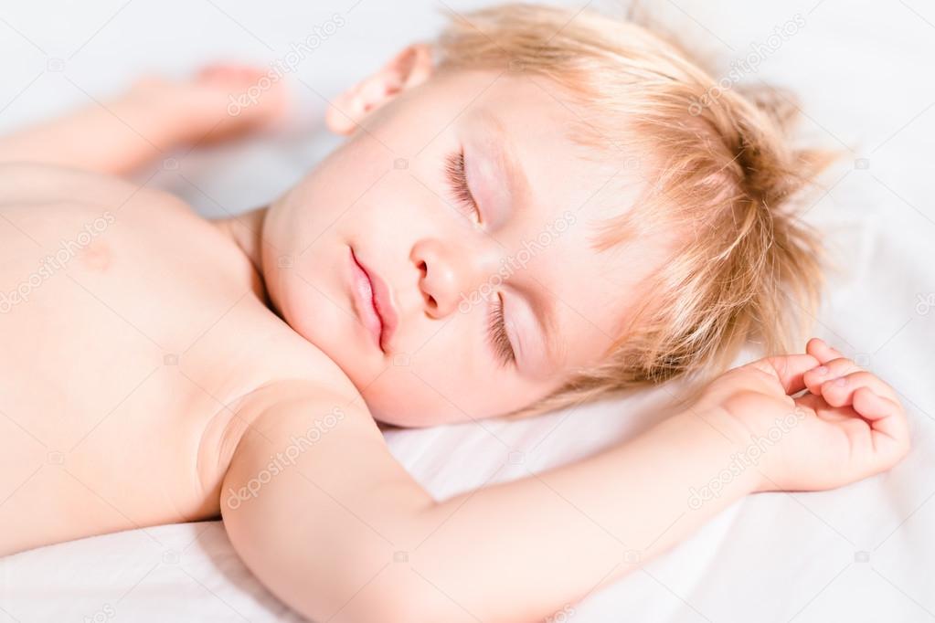 Close-up portrait of handsome toddler boy with blond hair sleeping on white bad. Carefree childhood concept