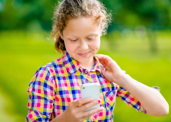 Adorable adolescente sonriente en ropa casual con teléfono inteligente en la mano, mirando a la pantalla, leyendo un mensaje, utilizando Facebook en el día soleado en el parque de verano Fotos de stock libres de derechos