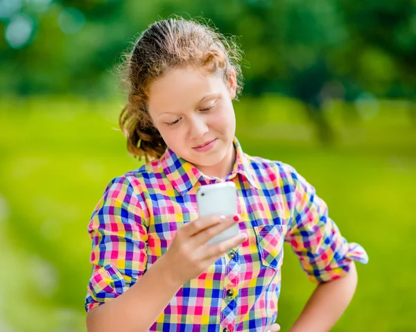 Досить посміхаючись Дівчинка-підліток в повсякденний одяг з смартфон в руці, дивлячись на екран, читання повідомлення, використовуючи Facebook в сонячний день в літній парк Стокова Картинка