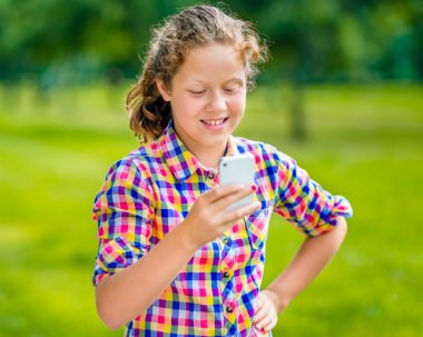 Facebook güneşli bir günde yaz parkta kullanarak bir ileti okuma ekranına, bakarak hoş gülümseyen genç kız rahat kıyafetler ile smartphone onun elinde