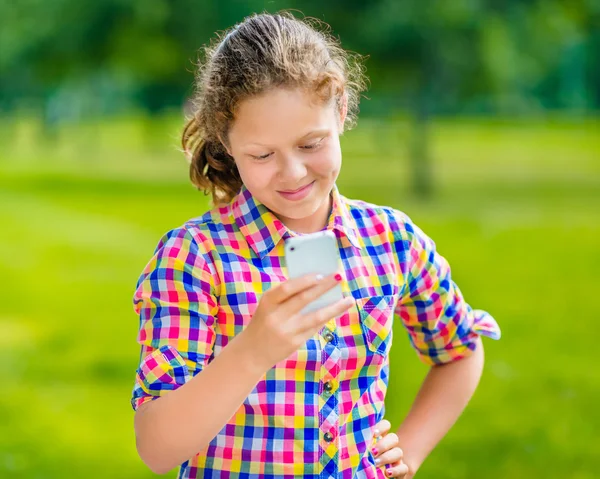 Dulce adolescente sonriente en ropa casual con teléfono inteligente en la mano, mirando a la pantalla, leyendo un mensaje, usando Facebook en el día soleado en el parque de verano Imagen de archivo