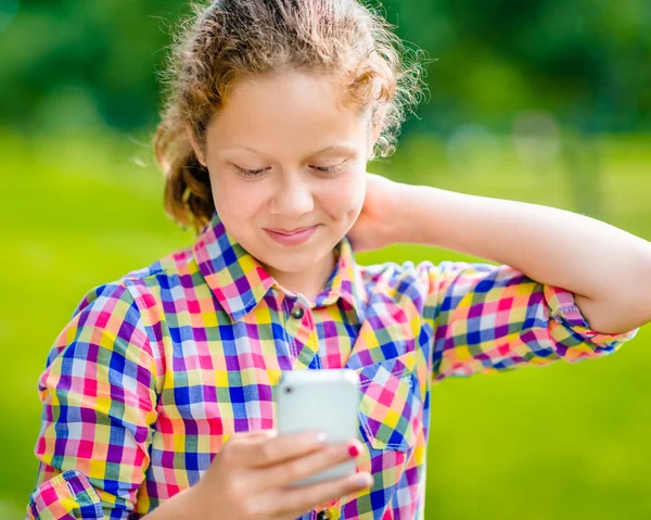 Hermosa adolescente sonriente en ropa casual con teléfono inteligente en la mano, mirando a la pantalla, leyendo un mensaje, usando Facebook en el día soleado en el parque de verano Fotos de stock
